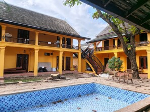 Un bâtiment jaune à deux étages, doté d’une piscine à carrelage bleu, d’un espace extérieur aménagé et d’un escalier menant à l’étage supérieur.