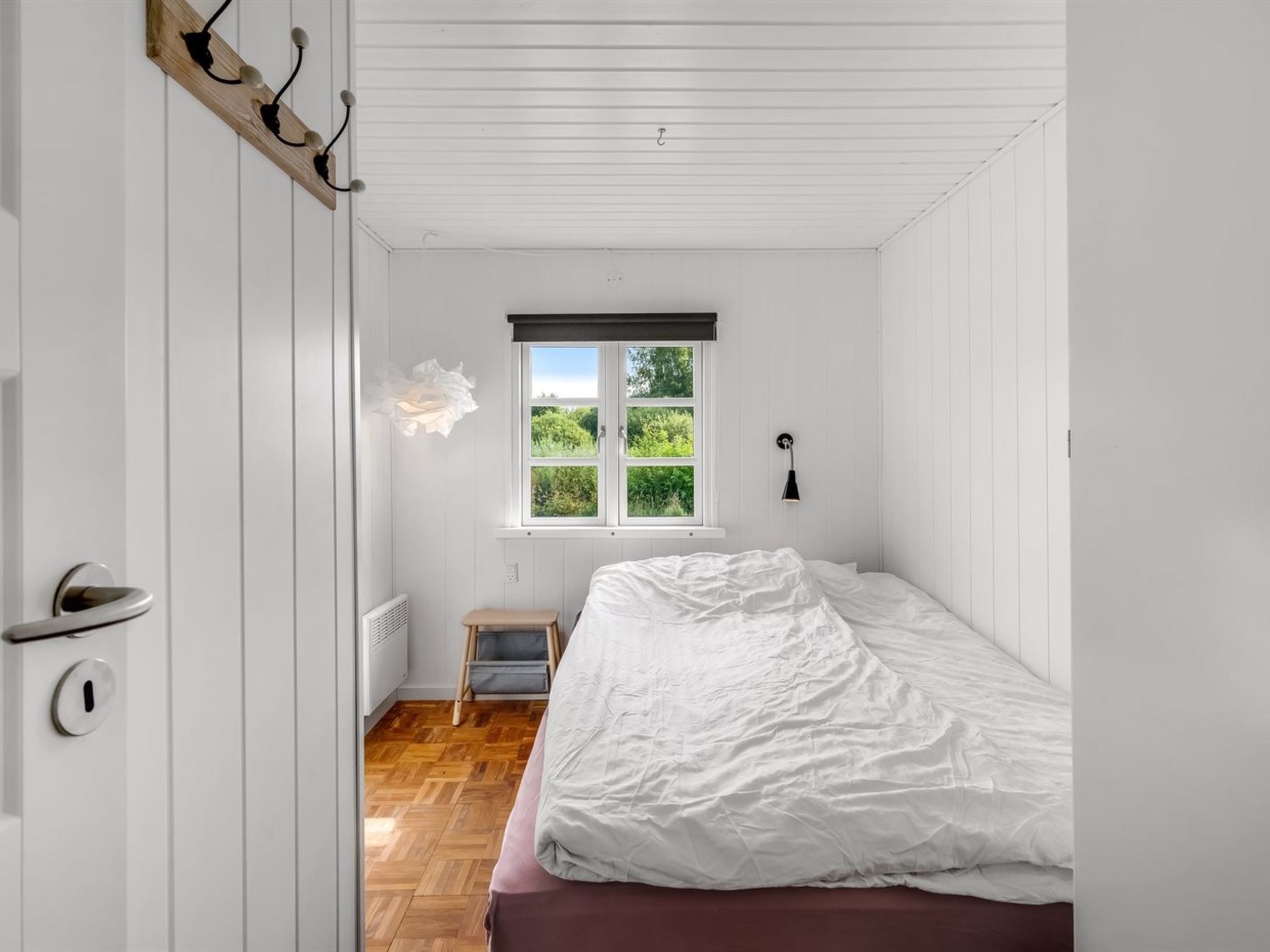 A bedroom with a bed, a window with a view of greenery, a radiator, and a door with a handle.