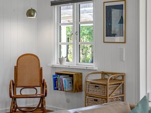 Um quarto com uma cadeira de vime, uma janela com vista, uma estante de livros e um quadro na parede.