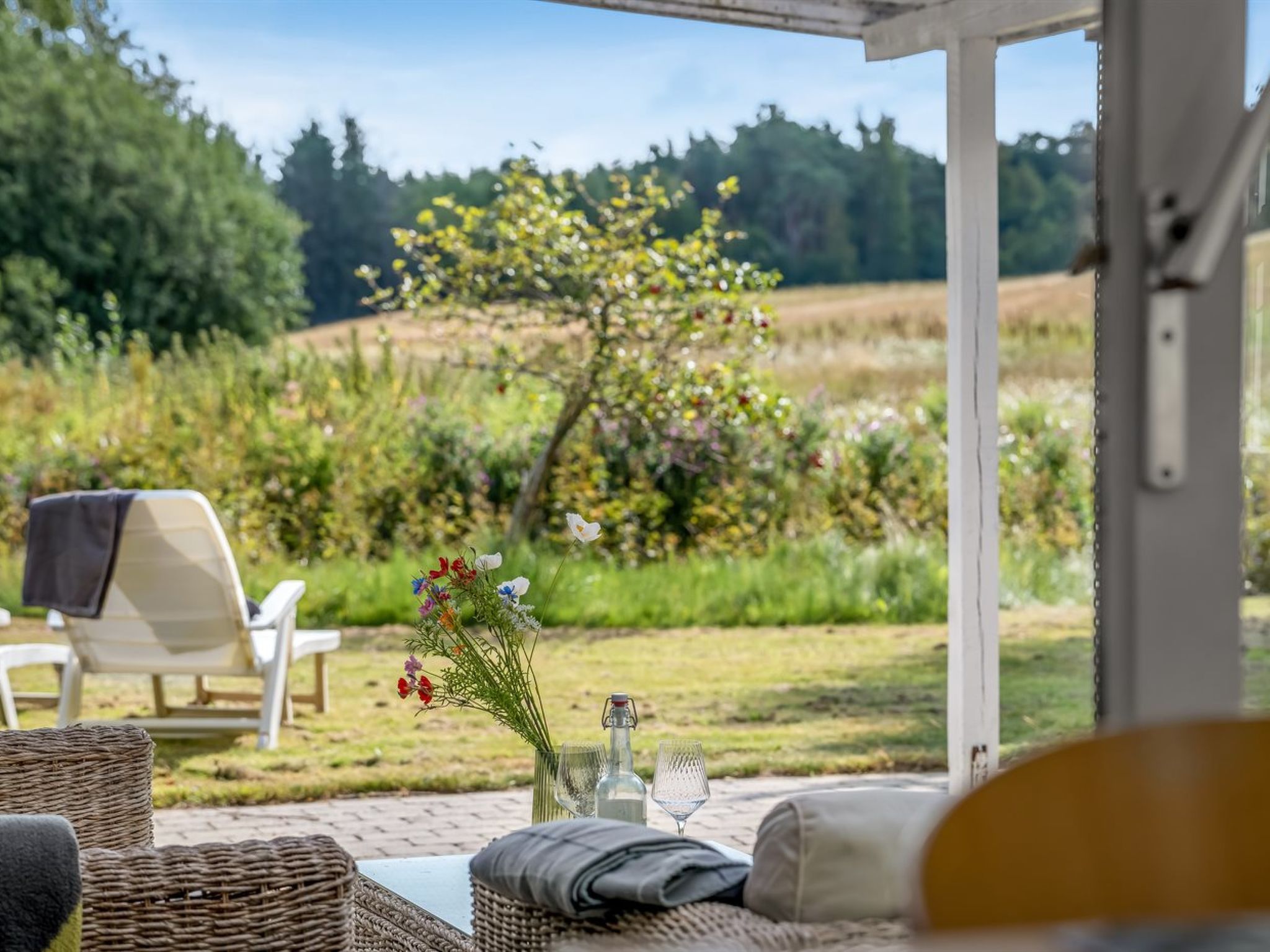 An outdoor seating area with wicker furniture, a table with a vase of flowers, and a view of a garden and fields.