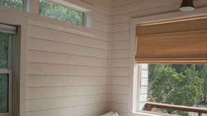 Quarto com cama, duas janelas e vista para árvores.