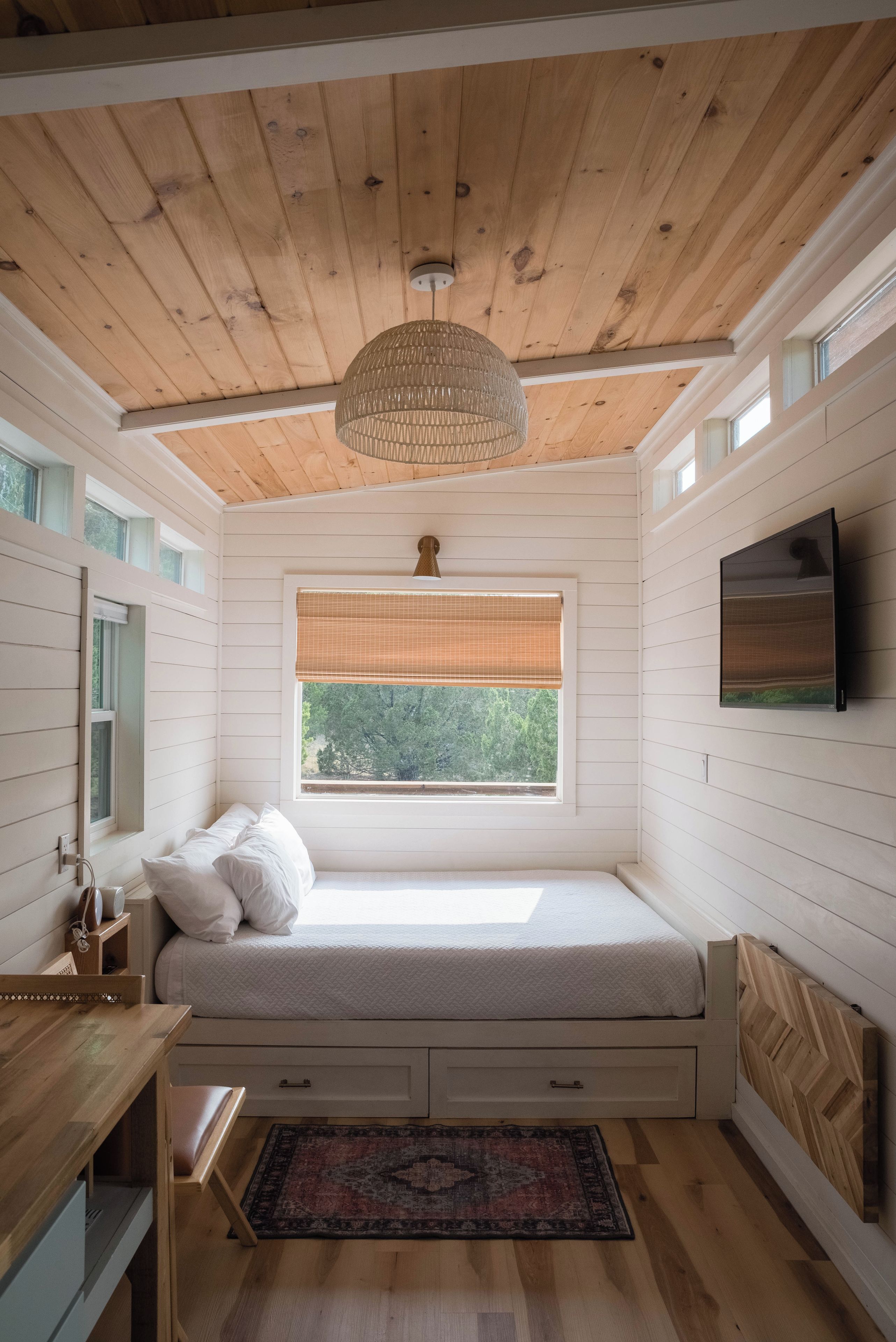 Un dormitorio con techo de madera, una cama con ropa blanca, una ventana con vistas a árboles, un televisor en la pared y una pequeña mesa de madera.