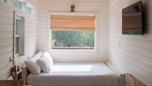 Quarto com teto de madeira, cama com roupa de cama branca, janela com vista para árvores, televisão na parede e uma pequena mesa de madeira.