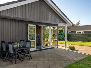 A covered patio with a table and chairs, a sliding glass door leading to a garden, and a wooden exterior wall. - "Fabian" - 300m from the sea by Interhome (Børkop)