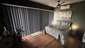 Un dormitorio con cama, ventilador de techo, mesita de noche con lámpara y ventana con persianas.