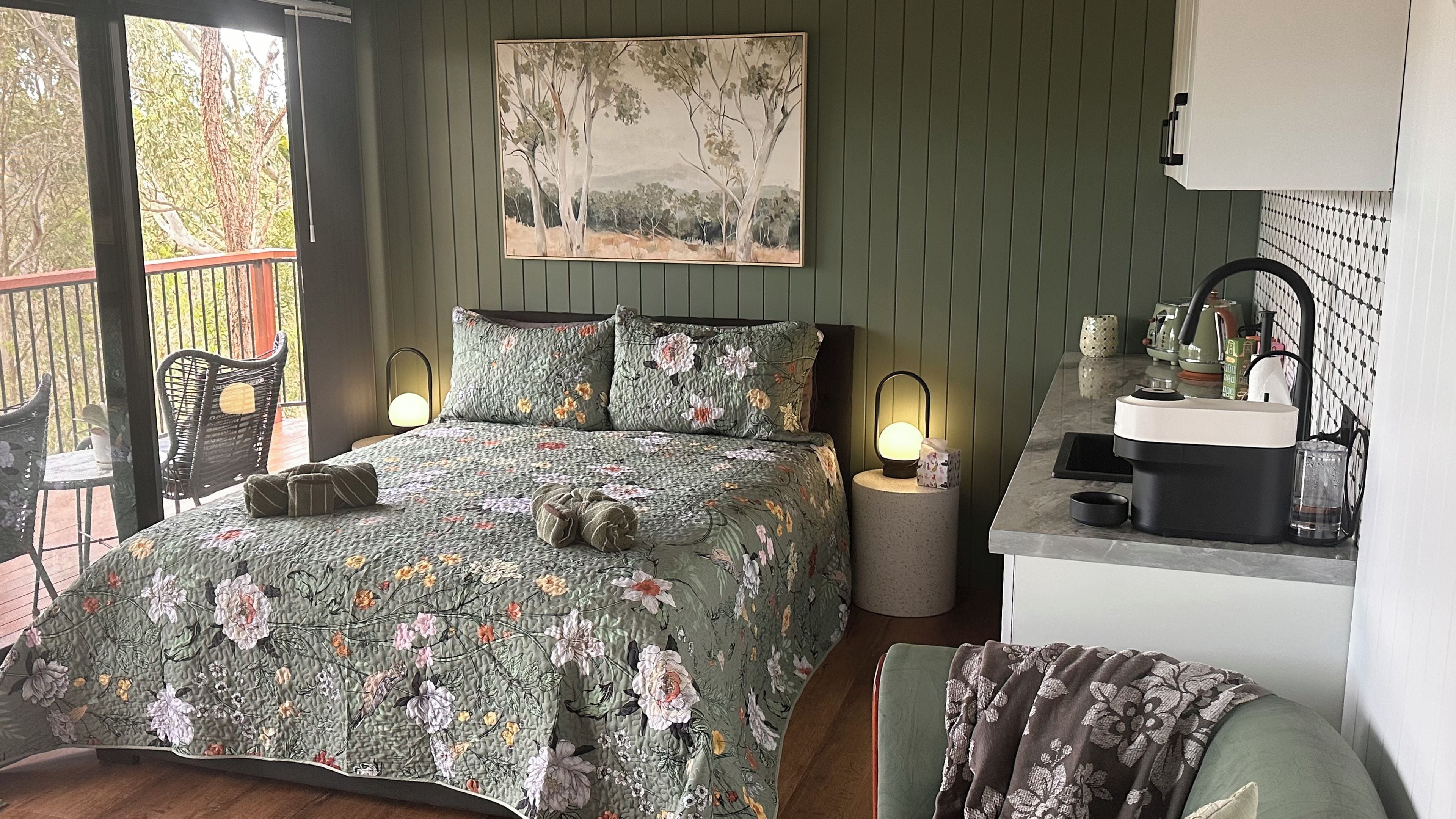 A bedroom with a bed, a painting, bedside lamps, a chair, and a small kitchenette.