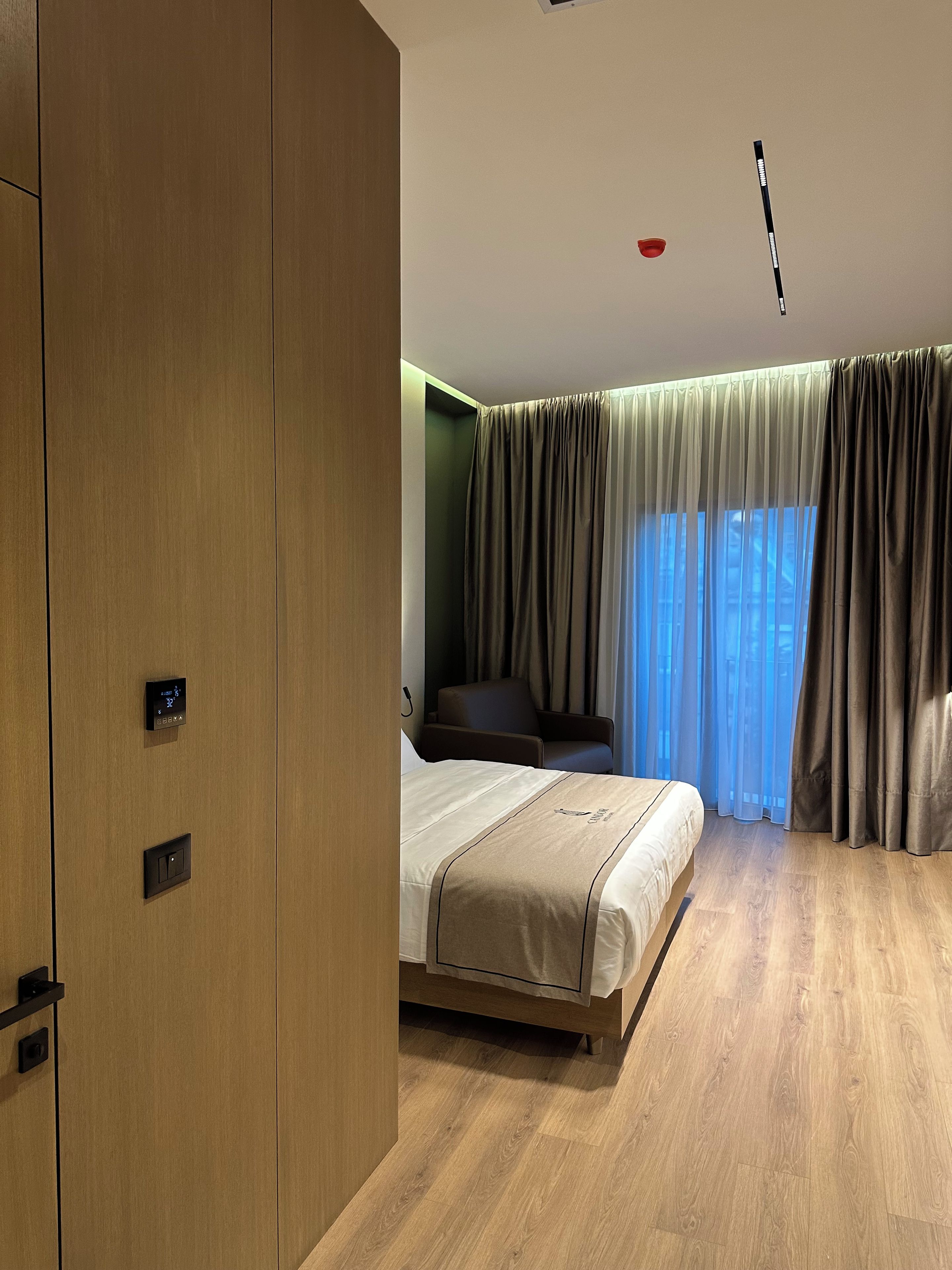 A modern hotel room with a wooden wardrobe, a bed with a beige bedspread, a seating area with a chair, and a large window with curtains.