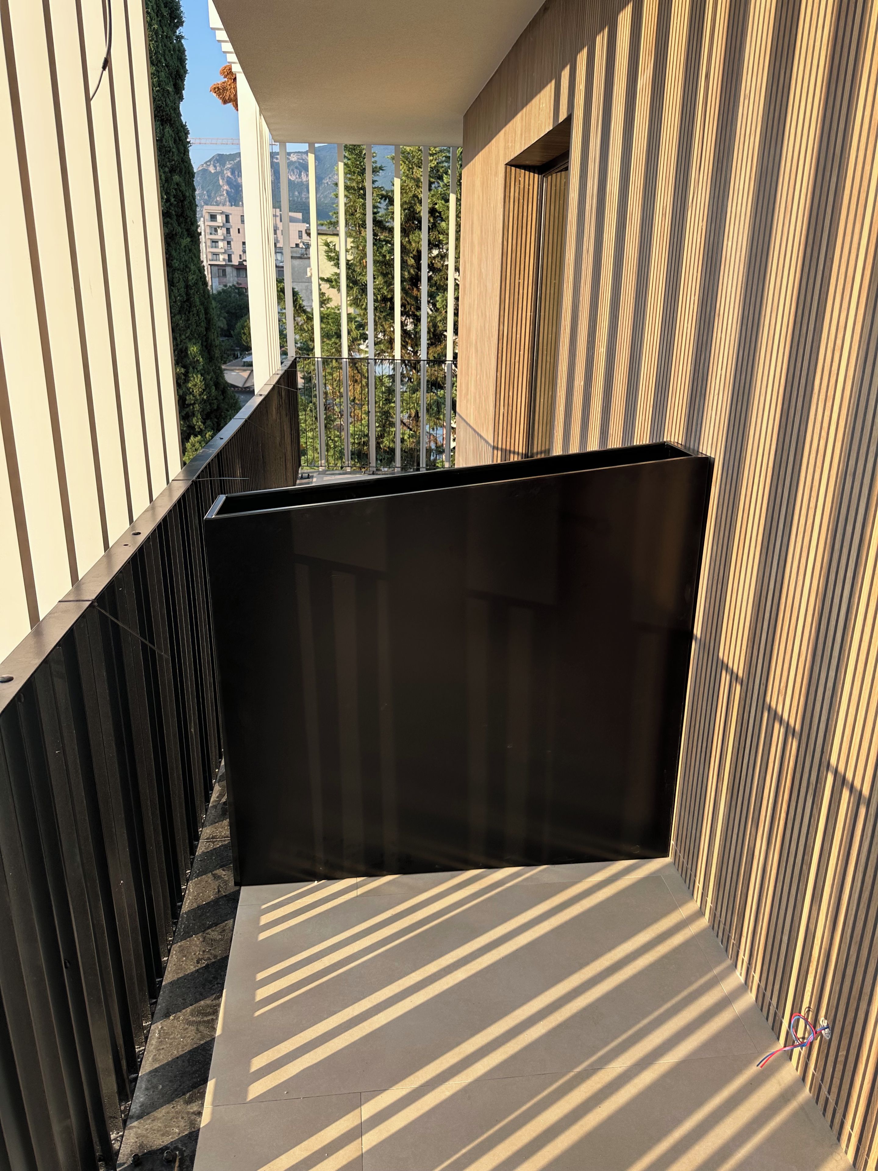 A balcony with a black trash bin, metal railings, and a view of buildings and trees.
