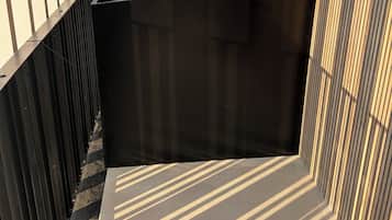 A balcony with a black trash bin, metal railings, and a view of buildings and trees.