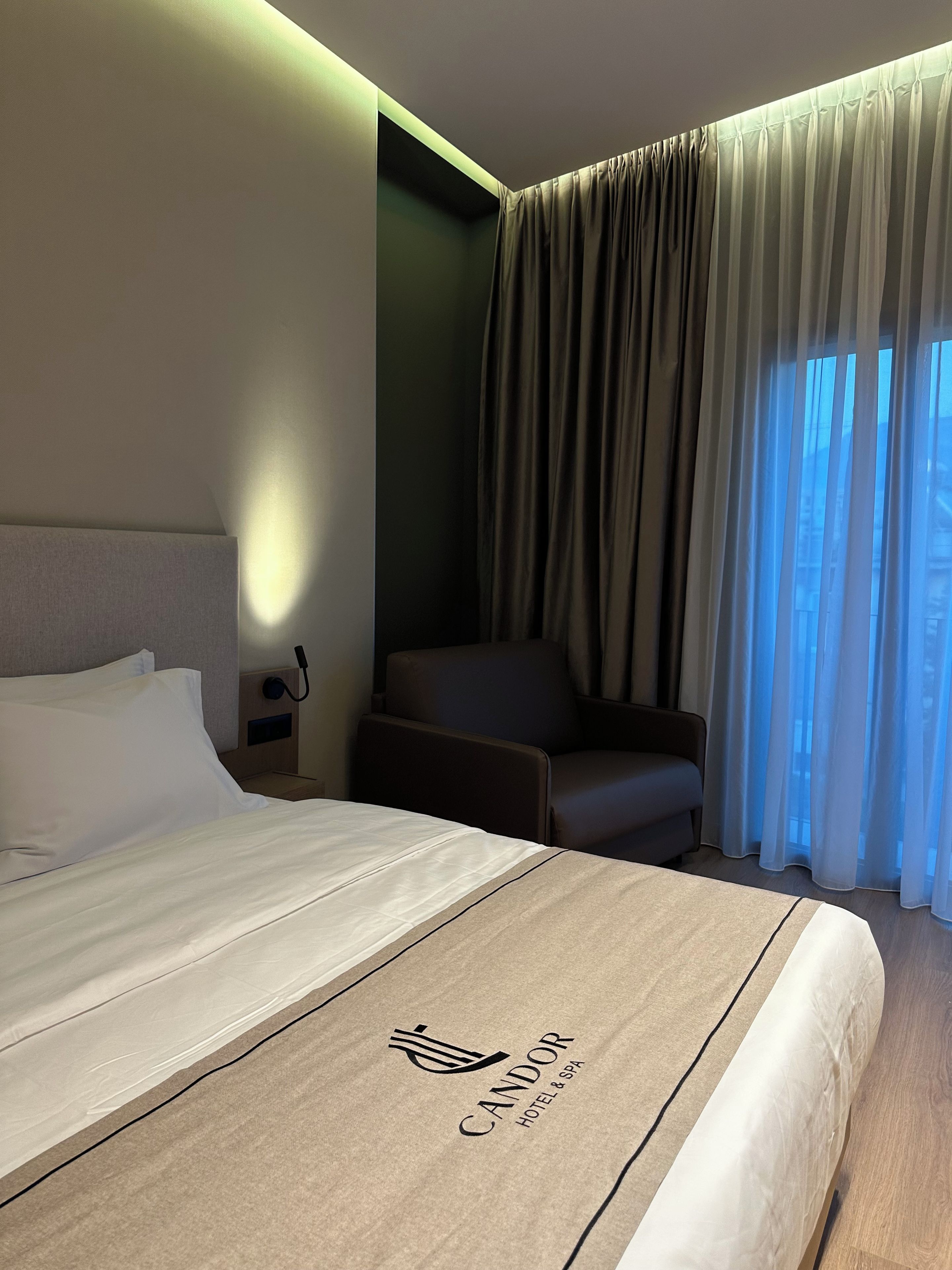 Ein Hotelzimmer mit Bett, Stuhl, Nachttisch und einem Fenster mit Vorhängen.