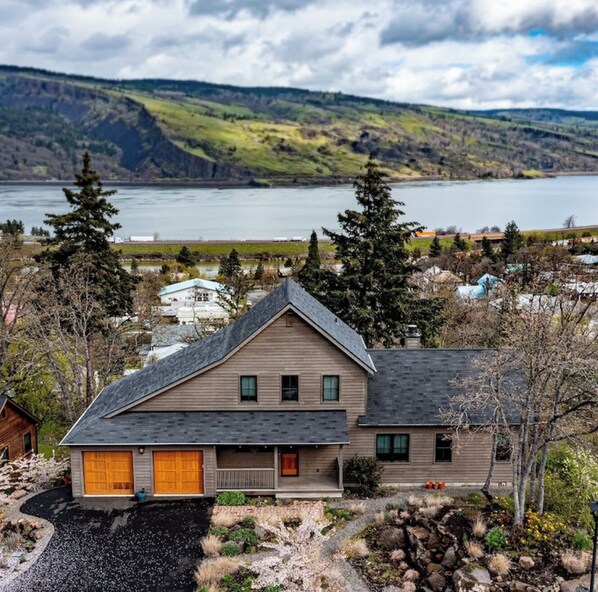 Uma casa de dois andares com uma garagem grande, cercada por um jardim bem cuidado e vista para um lago e colinas.