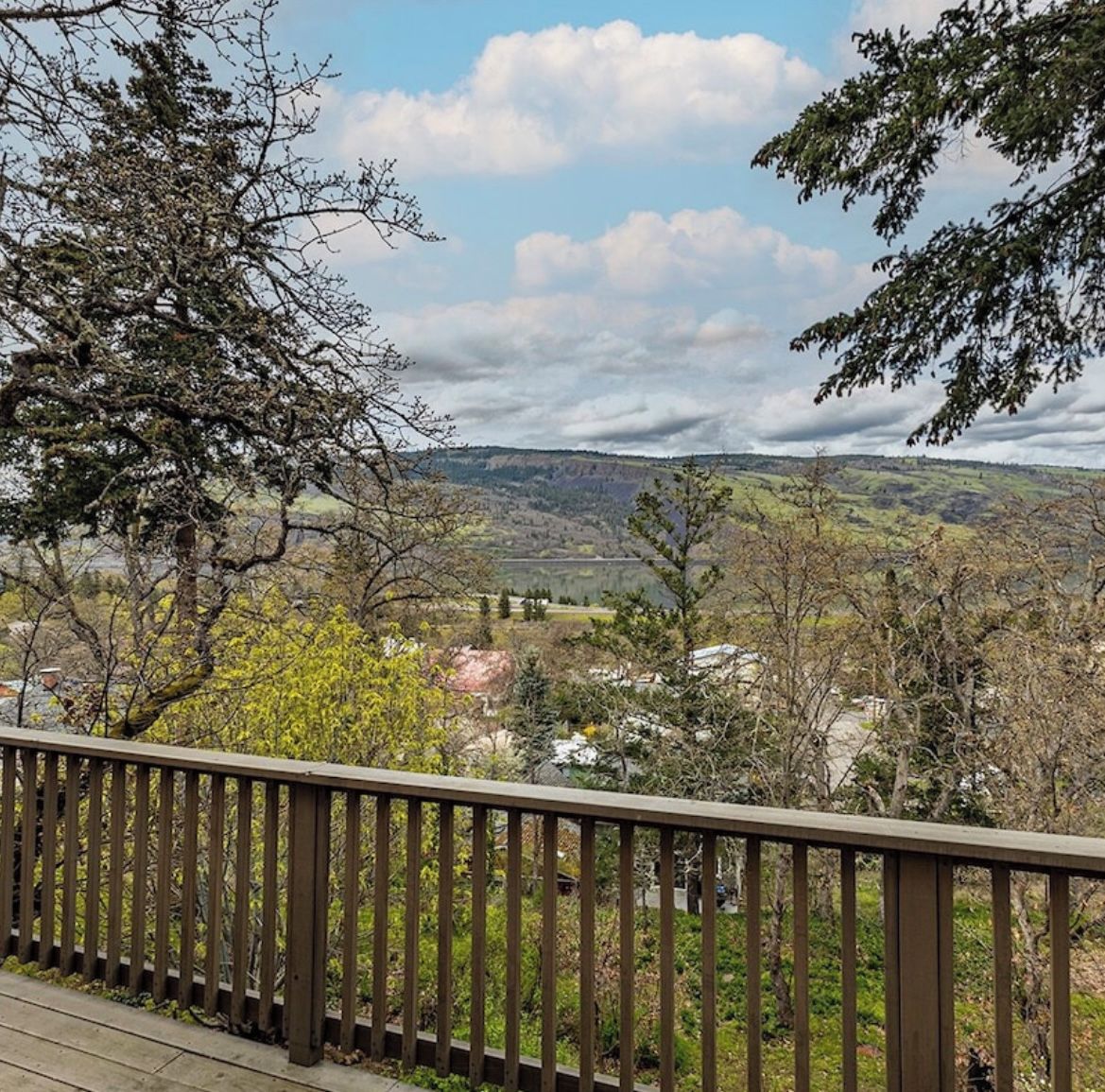 Blick von einem Balkon mit Holzgeländer auf ein Tal mit Bäumen und ferne Hügel.