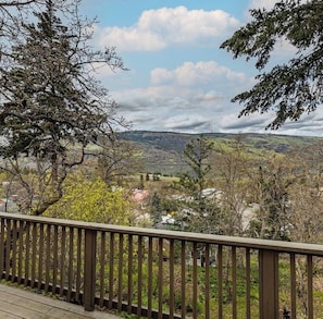 A view from a balcony with a wooden railing, overlooking a valley with trees and distant hills.