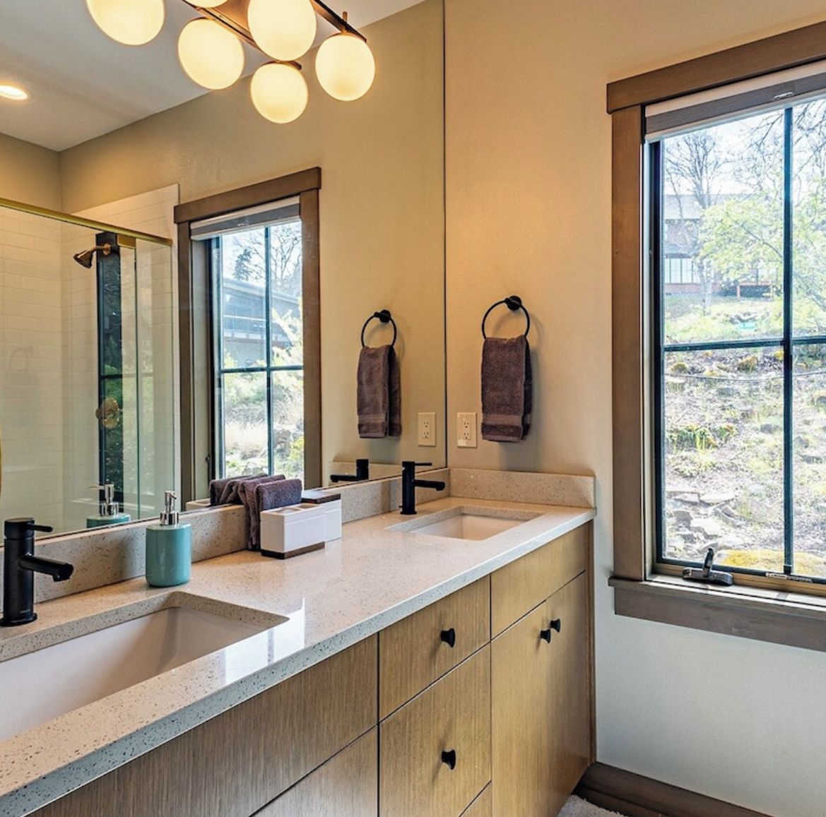 A modern bathroom with two sinks, a large mirror, and a window providing a view of the outdoors.