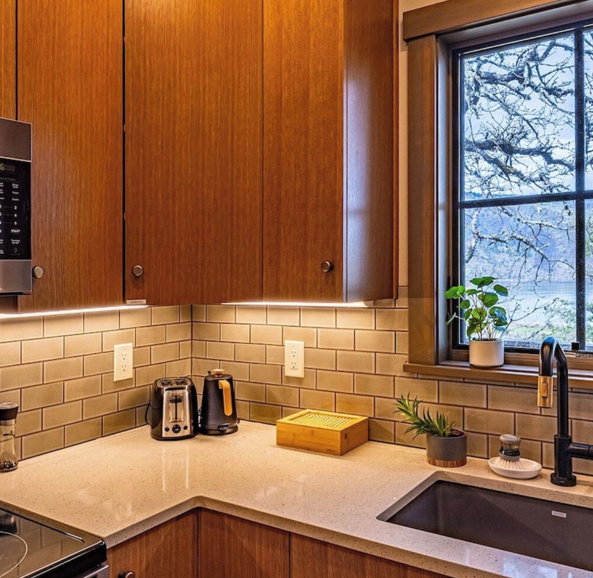 A kitchen with wooden cabinets, a countertop, a toaster, a coffee maker, and a window with a view of bare trees.
