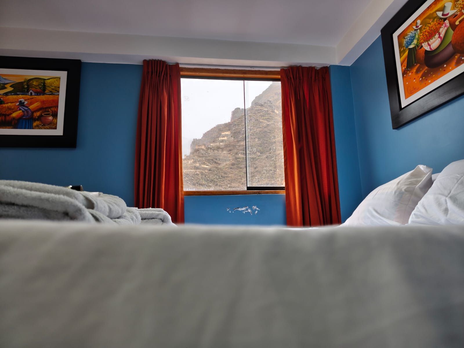 Una habitación de hotel con cama, ventana con cortinas y dos cuadros en la pared.