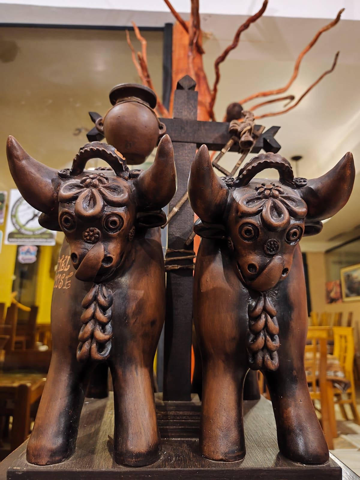 Eine Holzskulptur von zwei Ochsen, auf denen ein Topf steht, vor dem Hintergrund eines Baumes und eines Kreuzes.