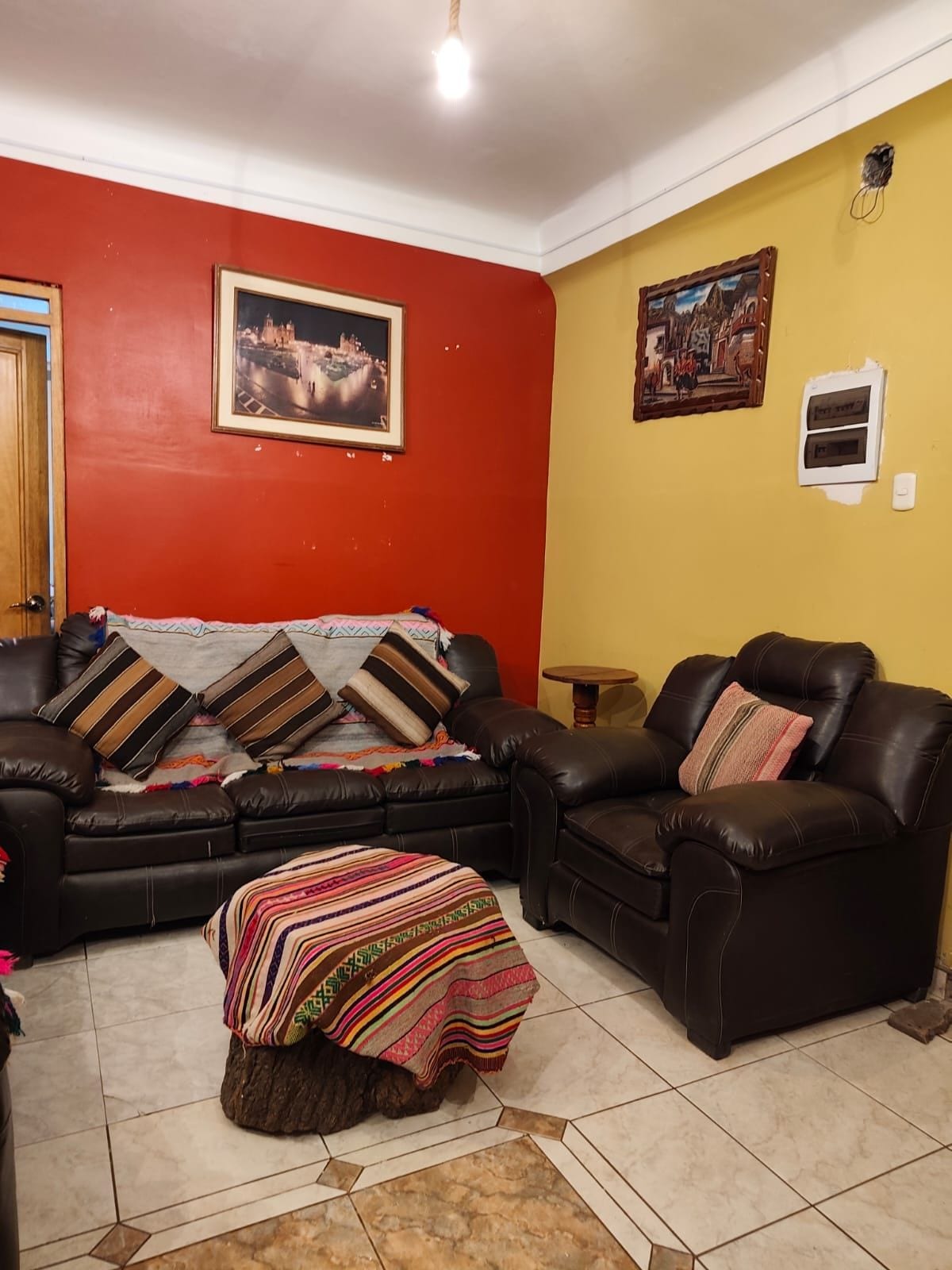 Uma sala de estar com sofá, duas poltronas, uma mesinha de madeira e um cobertor colorido no chão.