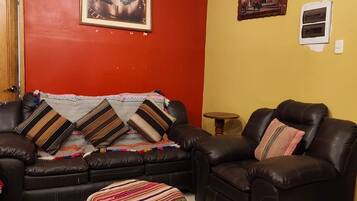 Uma sala de estar com sofá, duas poltronas, uma mesinha de madeira e um cobertor colorido no chão.