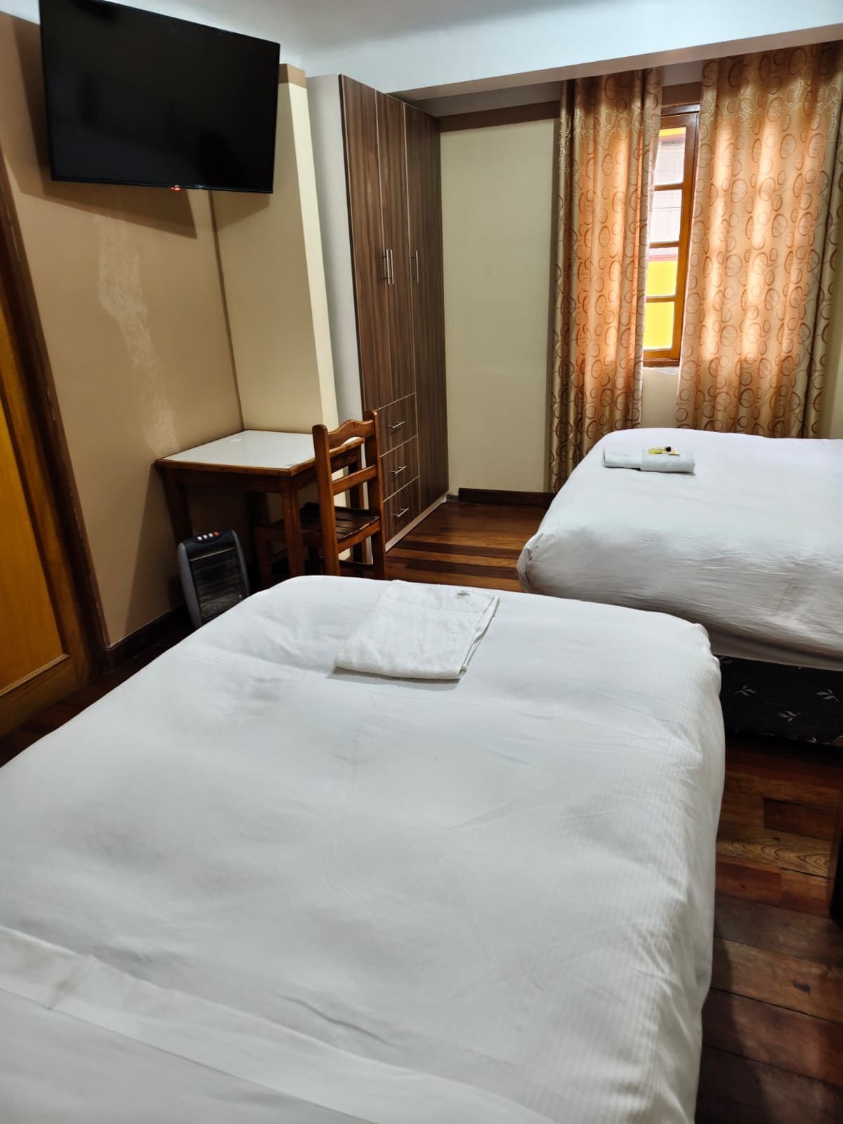 A hotel room with two beds, a desk, a chair, a TV mounted on the wall, and a window with curtains.