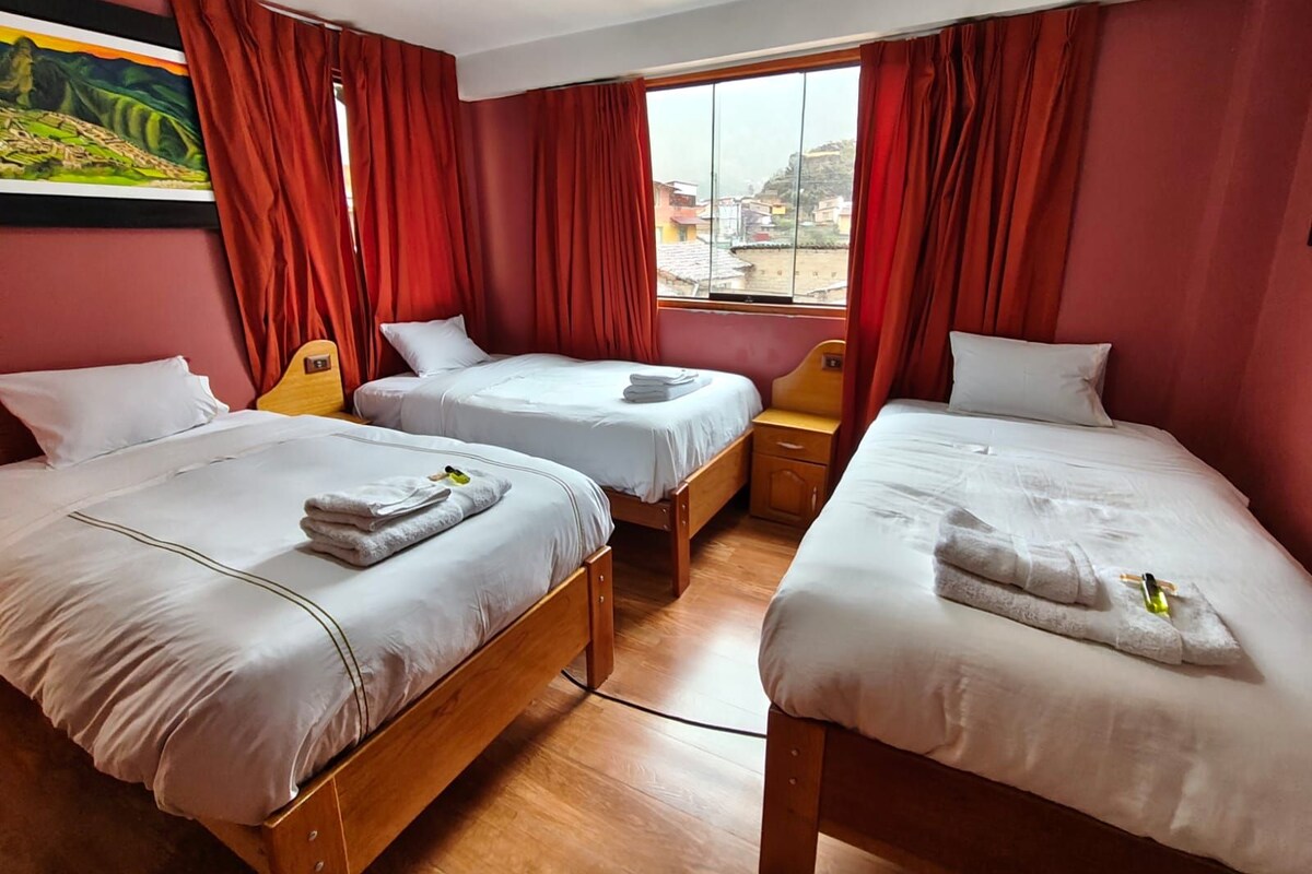 A hotel room with three beds, red curtains, and a window with a city view.