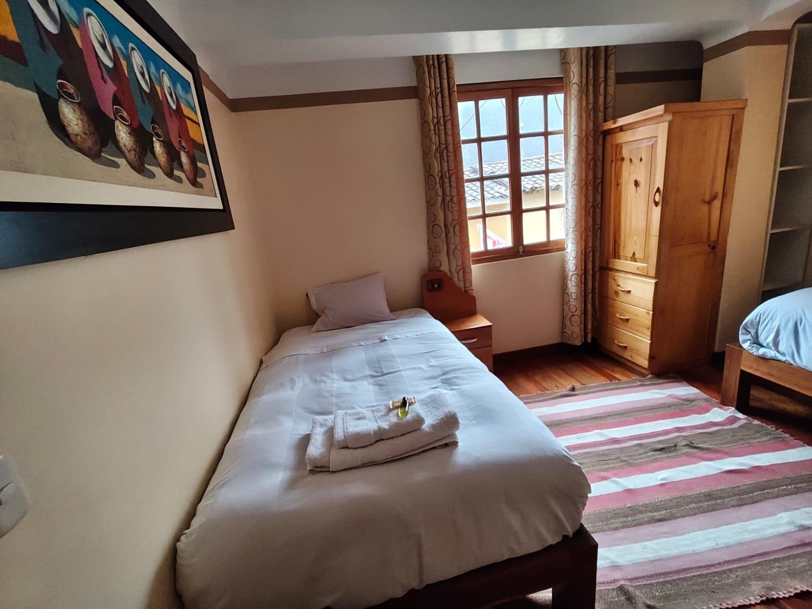A bedroom with a single bed, a wooden wardrobe, a window with curtains, and a painting on the wall.