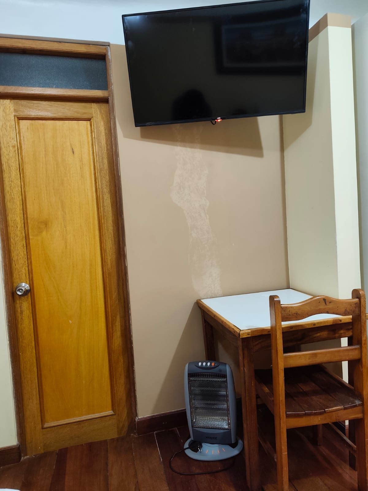 A room with a wooden door, a television mounted on the wall, a small table with a chair, and a portable heater.