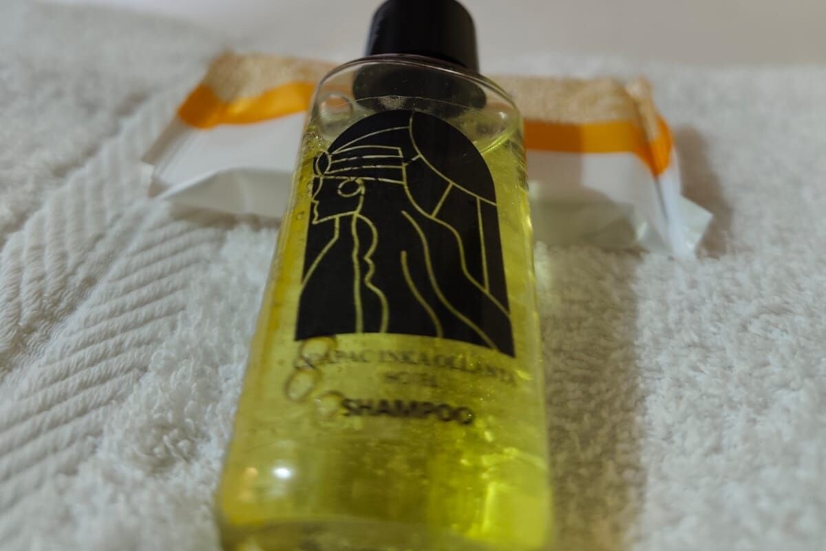 A bottle of shampoo on a bed with a window and curtains in the background.