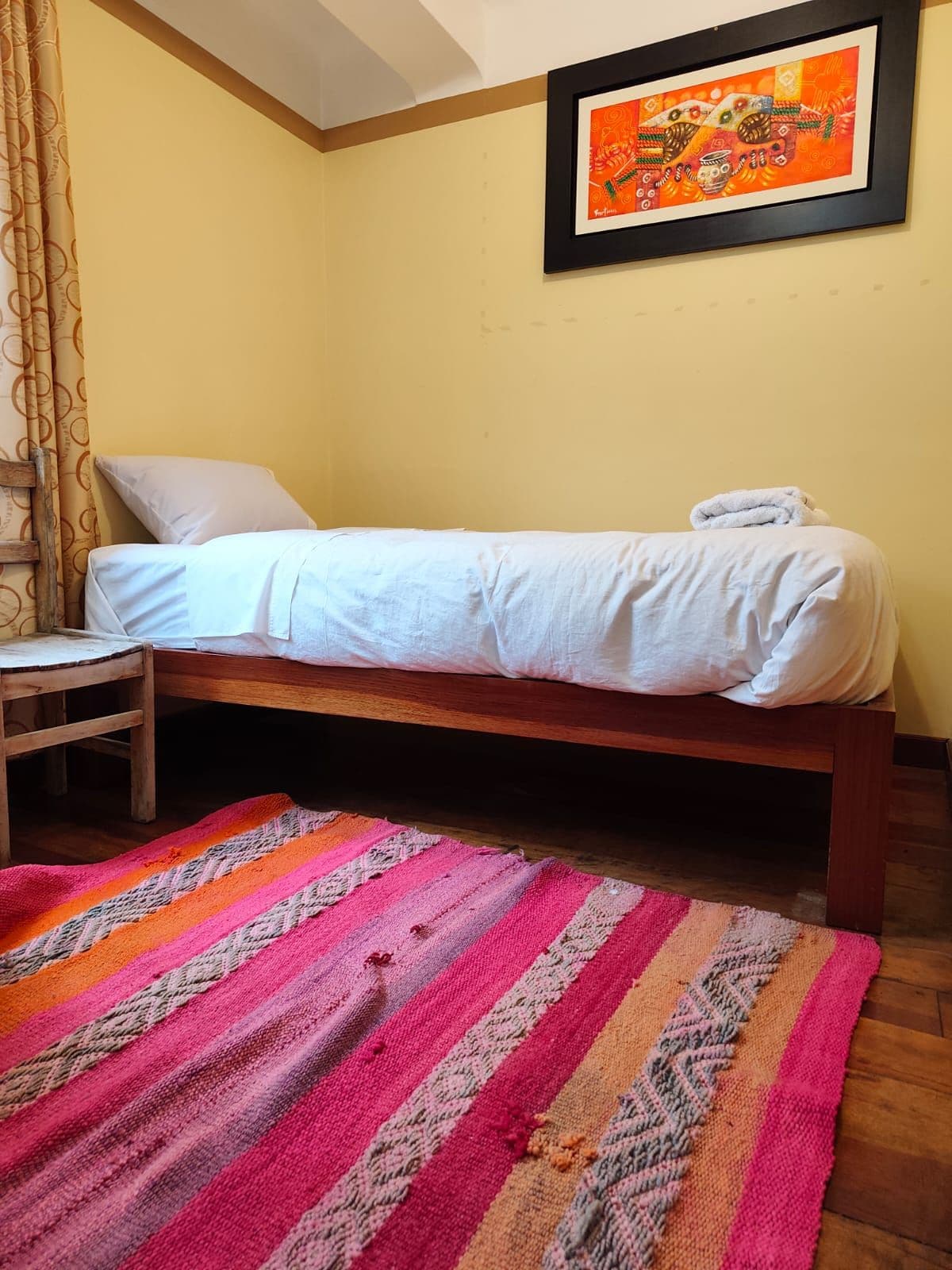 A single bed with a white sheet, a wooden headboard, and a colorful striped rug on the floor.