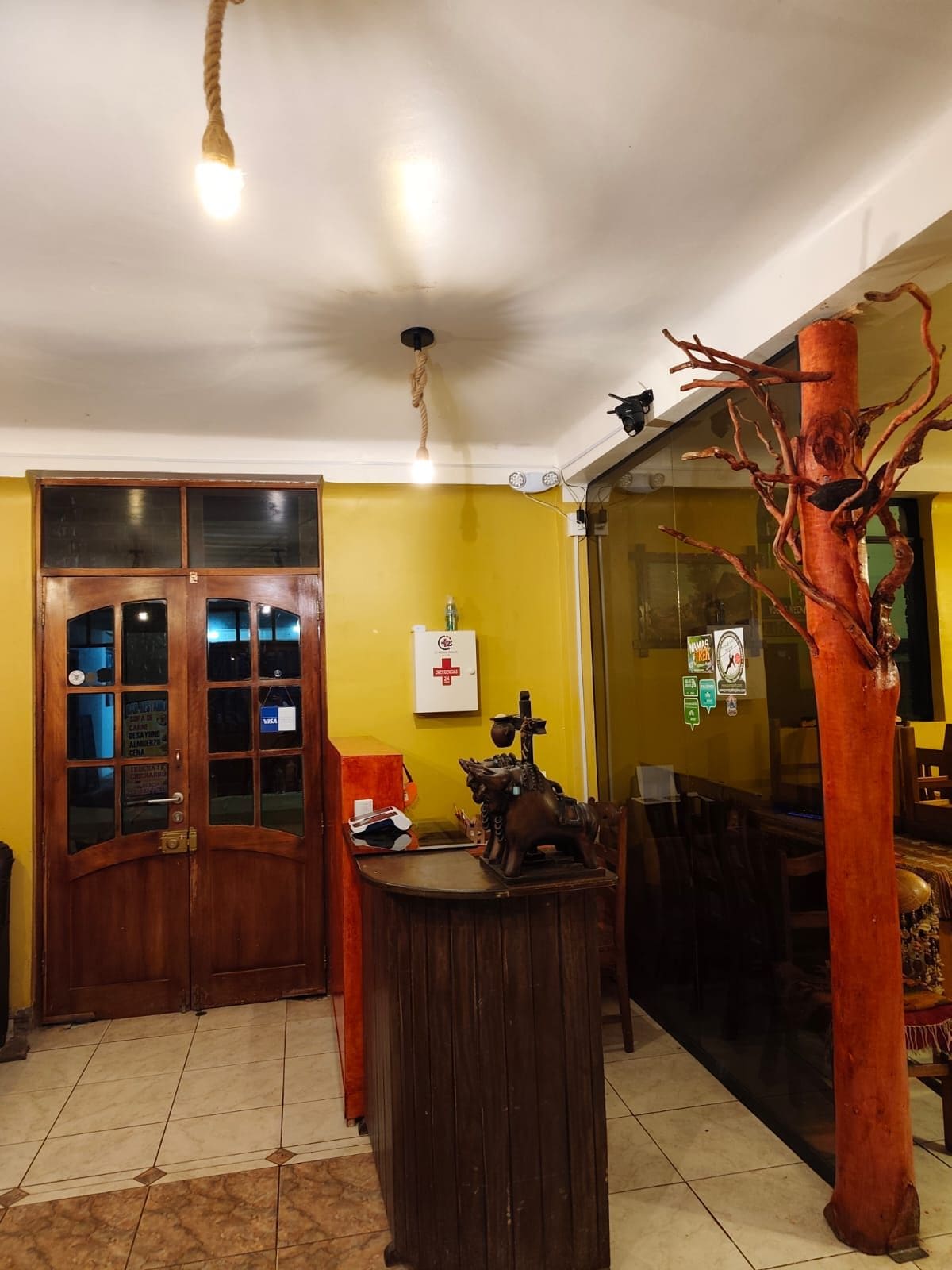 Quarto com porta de madeira, mesa redonda com um objeto decorativo, ventoinha no teto e luminária fixada na parede.