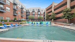A swimming pool with blue lounge chairs, surrounded by brick buildings with balconies. - King Suite | Walk to Toyota Stadium | Pool + Gym (Frisco)