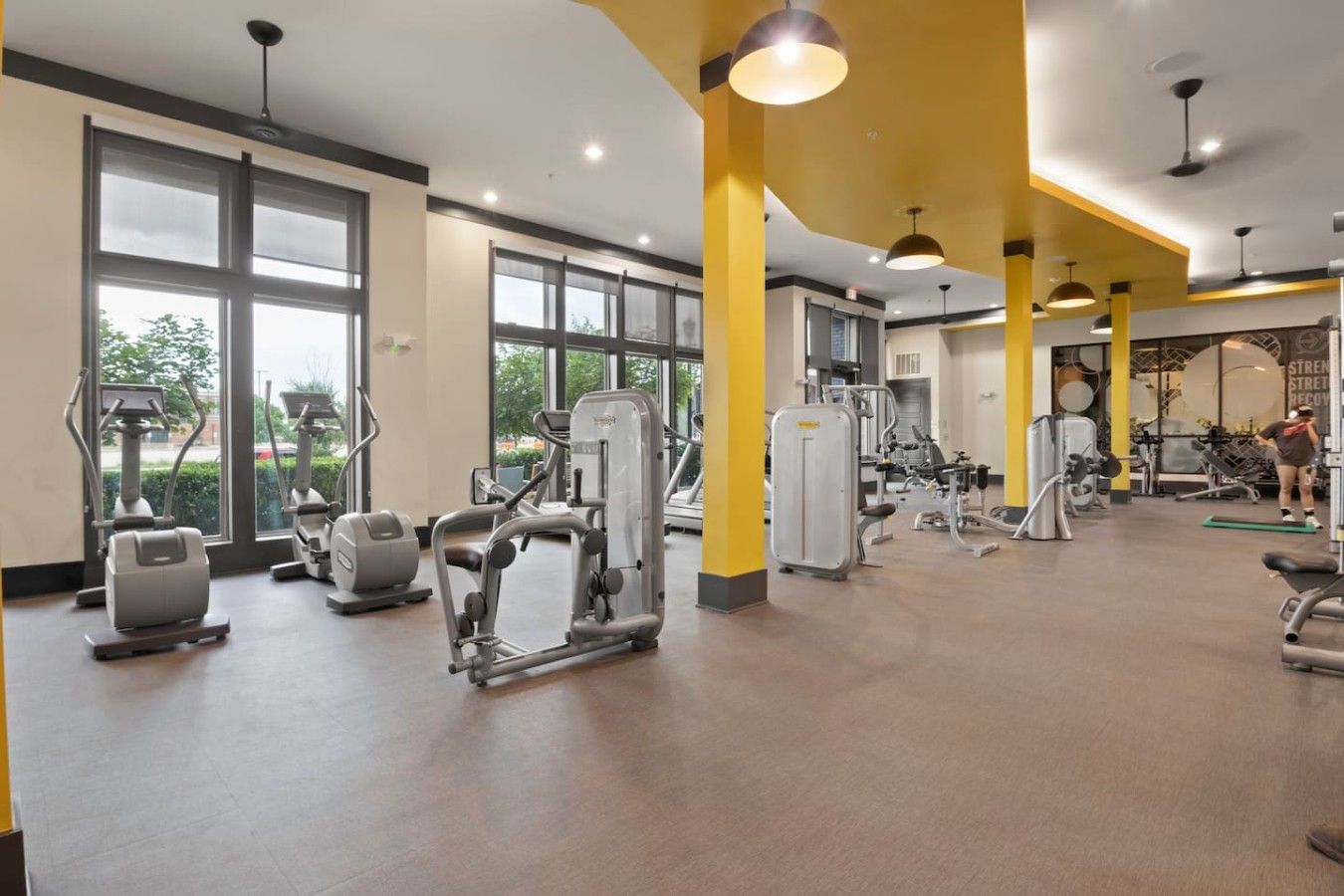 A gym with various exercise machines, large windows, and a yellow accent wall.