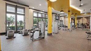 A gym with various exercise machines, large windows, and a yellow accent wall. - King Suite | Walk to Toyota Stadium | Pool + Gym (Frisco)