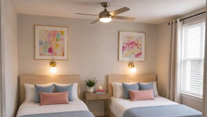 A bedroom with two beds, a ceiling fan, wall art, bedside lamps, and a window with curtains. - King Suite | Walk to Toyota Stadium | Pool + Gym (Frisco)