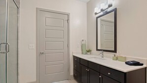 A bathroom with a white door, a glass shower enclosure, a dark vanity with a mirror, and a white sink. - King Suite | Walk to Toyota Stadium | Pool + Gym (Frisco)