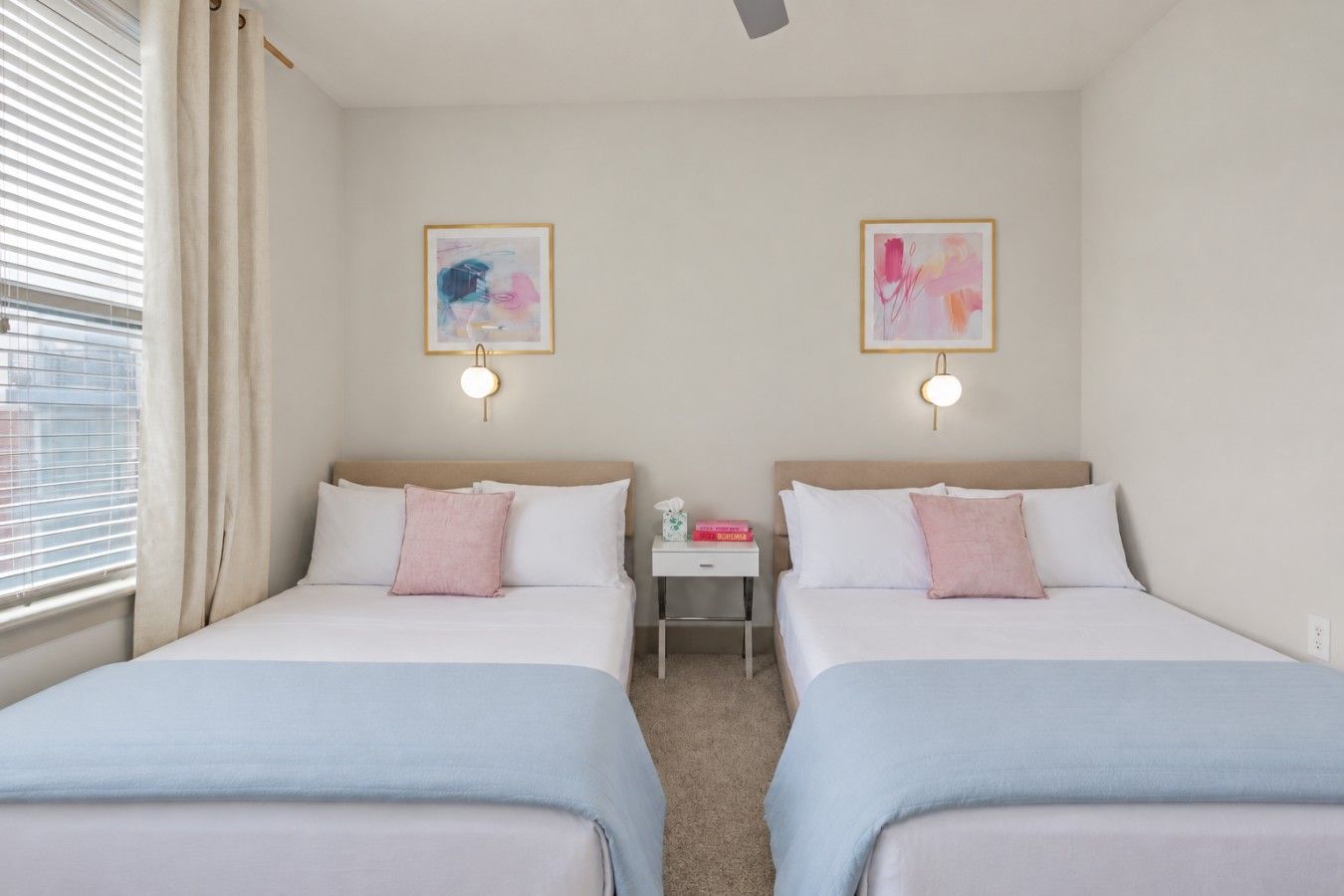 Ein Hotelzimmer mit zwei Betten, jeweils mit einem Kopfteil, einem Nachttisch mit Lampe sowie zwei gerahmten abstrakten Bildern an der Wand.