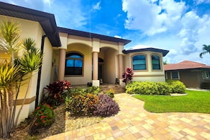 A residential house with a paved driveway, a covered porch, and a well-maintained garden with various plants and flowers. - Waterfront Fun & Relaxation Awaits at This Gulf-Access Getaway - Malaluka Sunset Villa - Roelens (Cape Coral)
