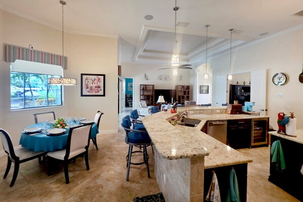 A spacious kitchen with a marble countertop, a dining area with blue tablecloths, and a view of a tropical landscape. - Waterfront Fun & Relaxation Awaits at This Gulf-Access Getaway - Malaluka Sunset Villa - Roelens (Cape Coral)