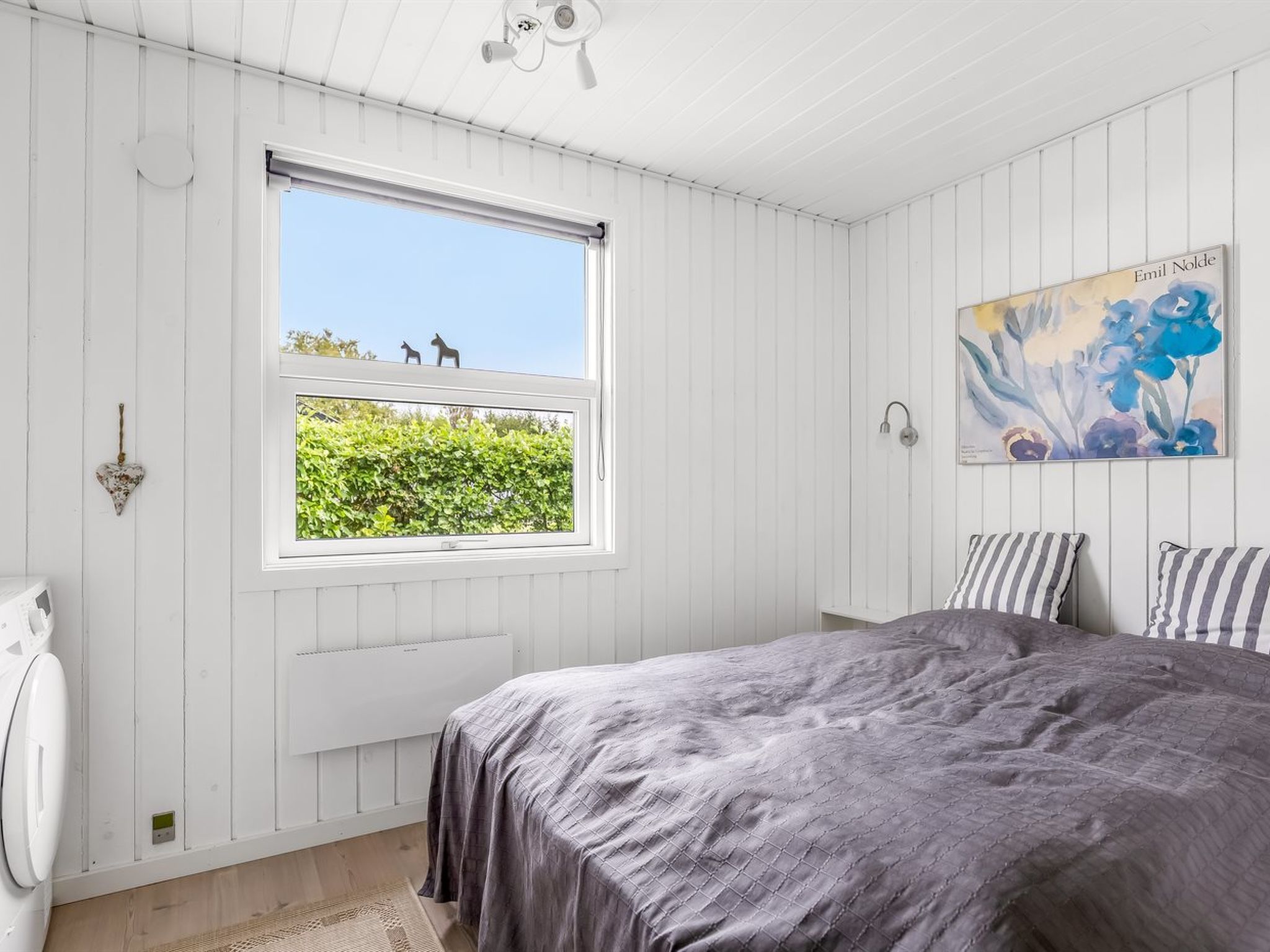 Une chambre avec une grande fenêtre, un lit recouvert d’une couette grise, un radiateur, un autre radiateur blanc, une lampe fixée au mur et un tableau.