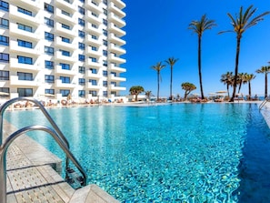 A swimming pool with clear blue water, surrounded by palm trees and a white building with balconies. - Ocean House Apartment Sea View Block 3, 7th Floor (Torremolinos)