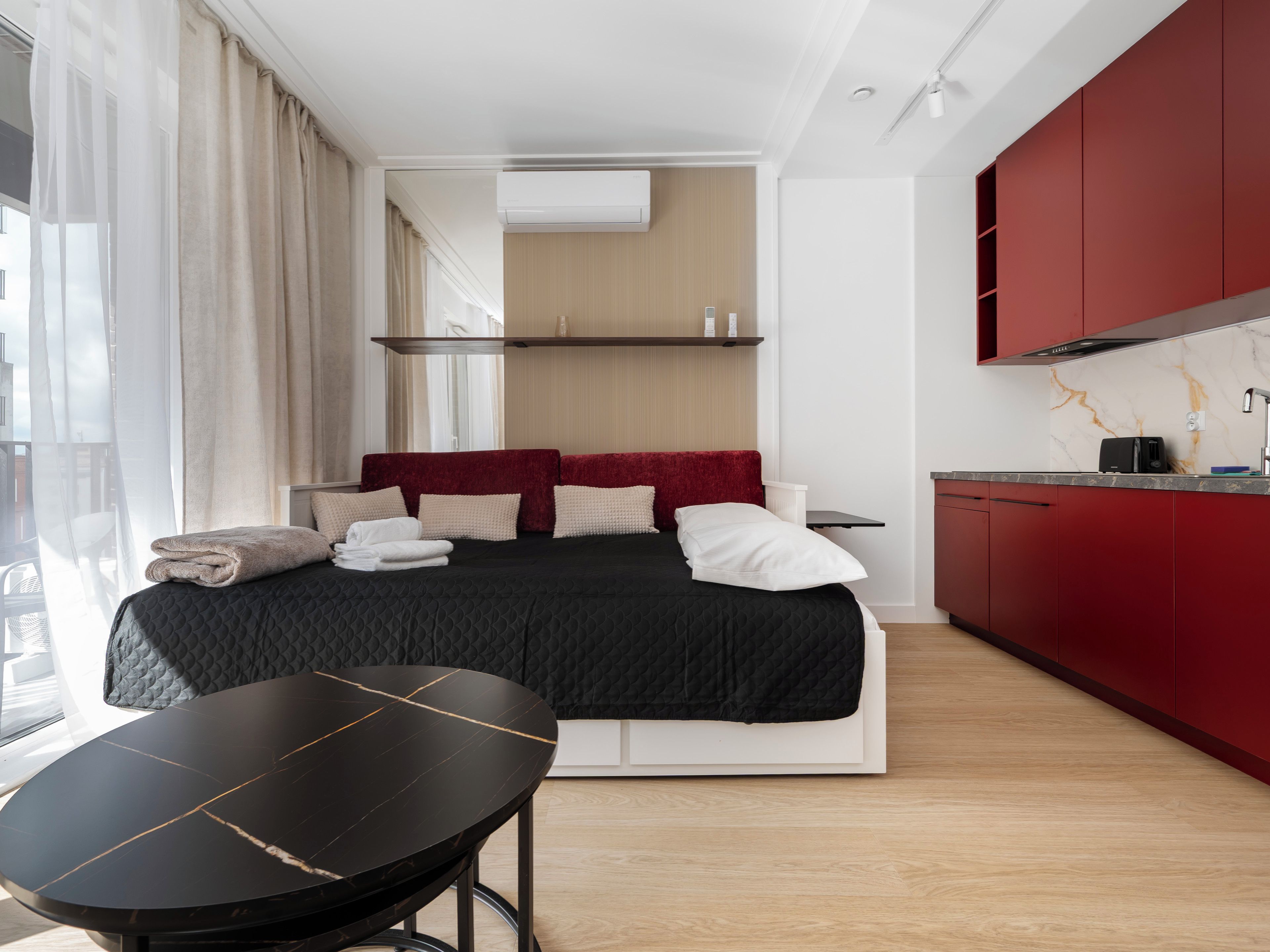 A modern hotel room with a bed, a dining table, a kitchen area, and a large window with curtains.