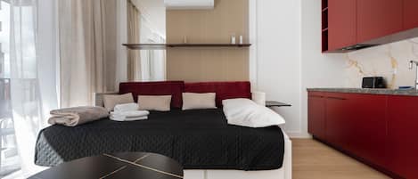 Una habitación de hotel moderna con cama, mesa de comedor, zona de cocina y un amplio ventanal con cortinas.
