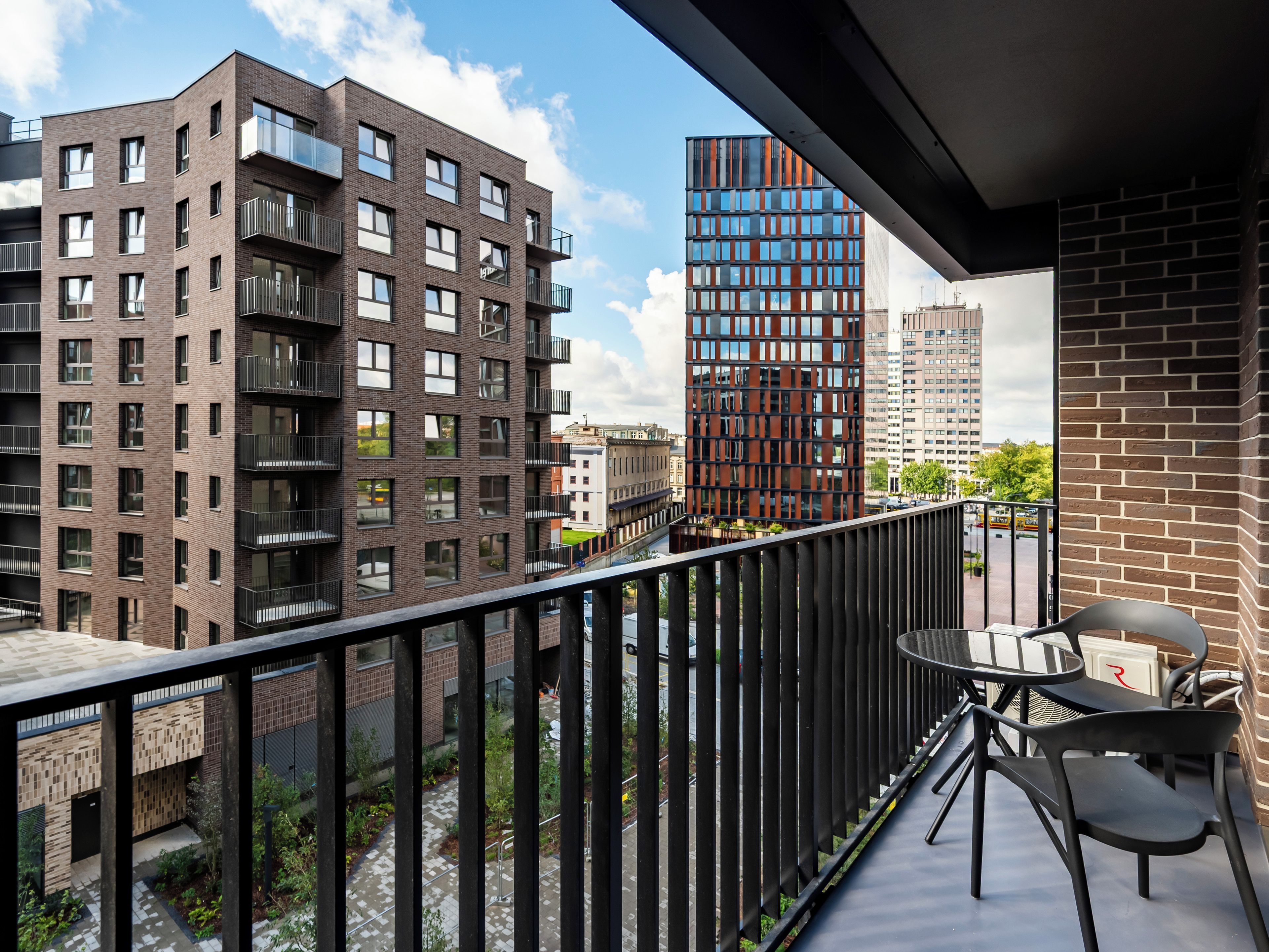 A balcony with a view of modern buildings, a table with chairs, and a small garden area.