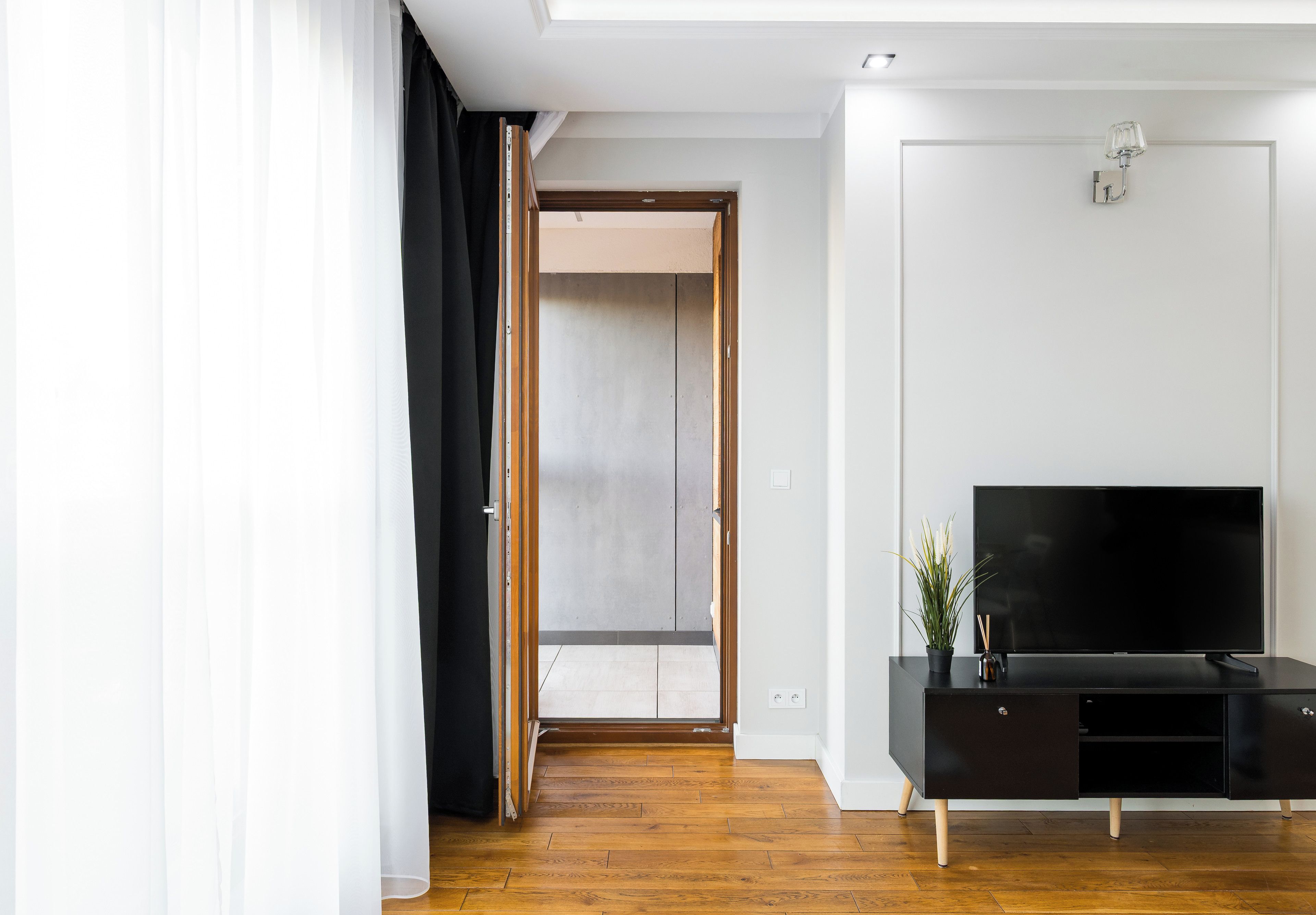 Una sala de estar moderna con piso de madera, un mueble negro para equipo audiovisual con televisor de pantalla plana, y una puerta que da a un balcón.