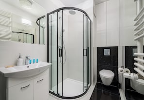 Un baño moderno con ducha envuelta en vidrio, un lavamanos blanco con artículos de aseo y un inodoro con dispensador de toallas de papel.