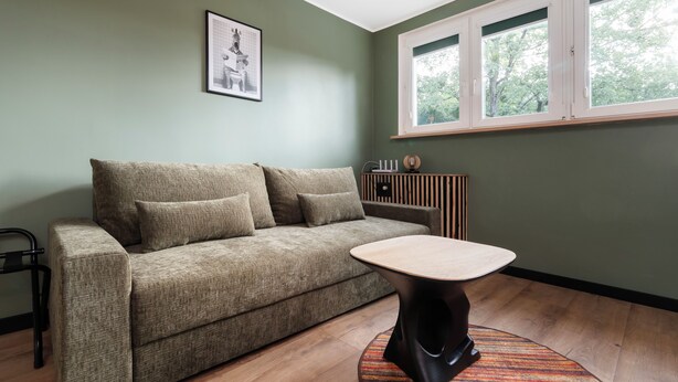 Una habitación con sofá, una mesa pequeña, un radiador y una ventana con vistas a árboles.