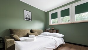 Un dormitorio con cama, almohadas, una silla, una mesita de noche, un cuadro enmarcado y una ventana.