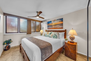 A bedroom with a bed, two bedside lamps, a ceiling fan, a window with blinds, a wall-mounted air conditioner, and a painting of a sunset. - Oceanfront Lahaina Oasis 1 Bedroom (Lahaina)