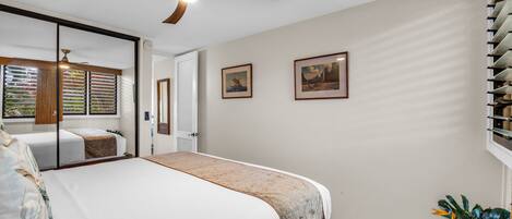 A bedroom with a bed, a ceiling fan, a window with blinds, and two framed pictures on the wall.