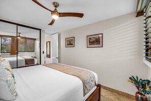 A bedroom with a bed, a ceiling fan, a window with blinds, and two framed pictures on the wall. - Oceanfront Lahaina Oasis 1 Bedroom (Lahaina)